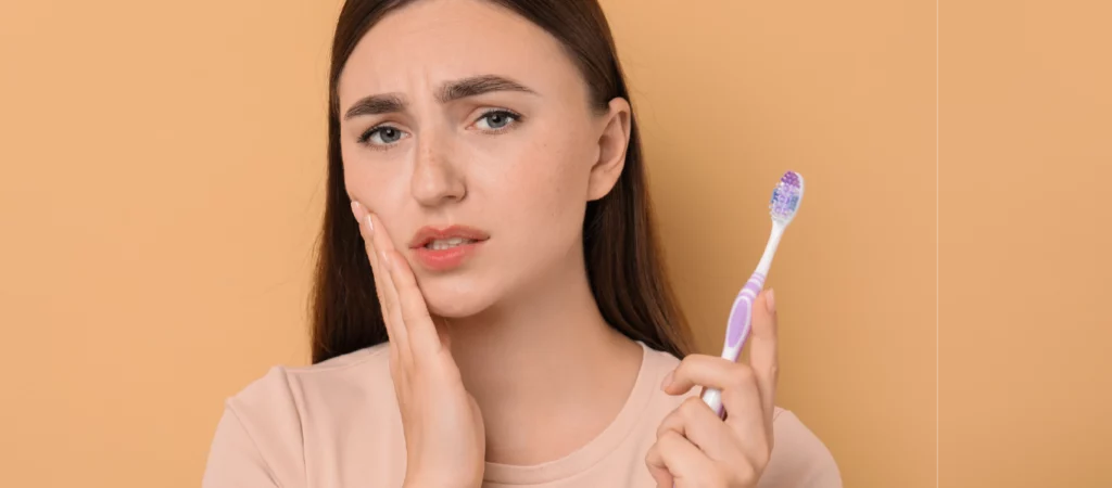 gengivas e hormonas: mulher com a mão na face e uma escova de dentes na mão